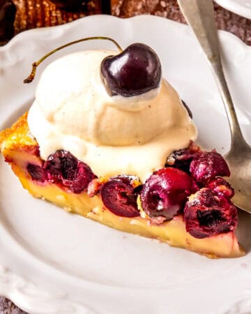 A slice of cherry clafoutis topped with creamy vanilla ice cream and a sweet cherry, served on a white plate with a silver spoon.