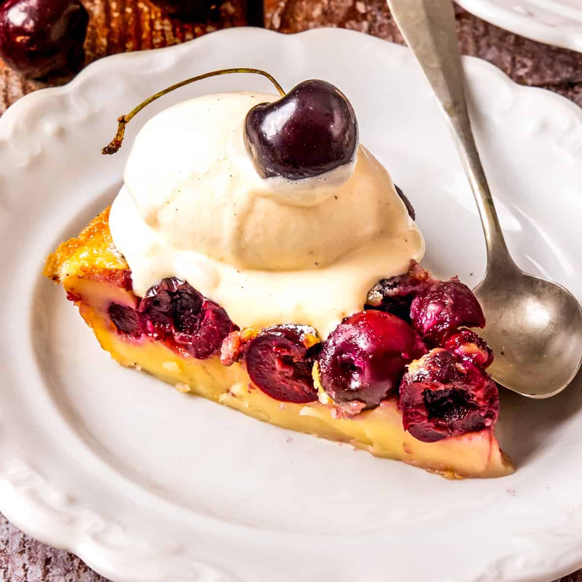 A slice of cherry clafoutis topped with creamy vanilla ice cream and a sweet cherry, served on a white plate with a silver spoon.