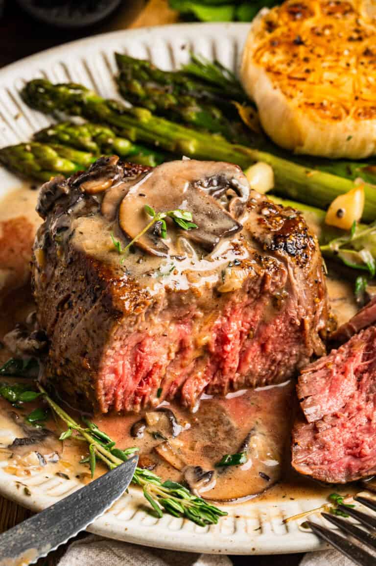 A juicy filet mignon topped with mushroom sauce is served with roasted asparagus and a halved head of garlic on a white plate. A slice of steak is cut and a knife and fork are placed nearby.