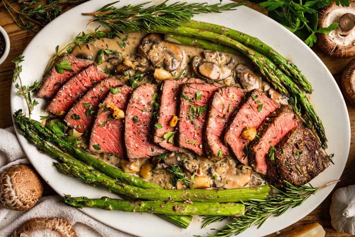 20 Best Easter Dinner Ideas (Main Dishes) Sliced medium-rare steak with a creamy mushroom sauce, garnished with herbs, served alongside roasted asparagus on a white oval platter. Fresh herbs and mushrooms are scattered around the dish.