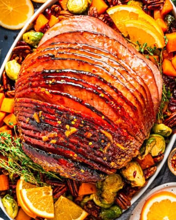 A glazed, spiral-cut ham garnished with thyme is served on a platter with roasted brussels sprouts, cubes of butternut squash, pecans, and orange slices. Small bowls of spices and orange wedges are nearby.