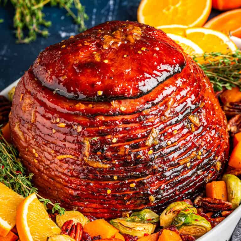 A glazed, spiral-cut baked ham sits on a platter, garnished with orange slices, roasted brussels sprouts, pecans, herbs, and chunks of roasted butternut squash.