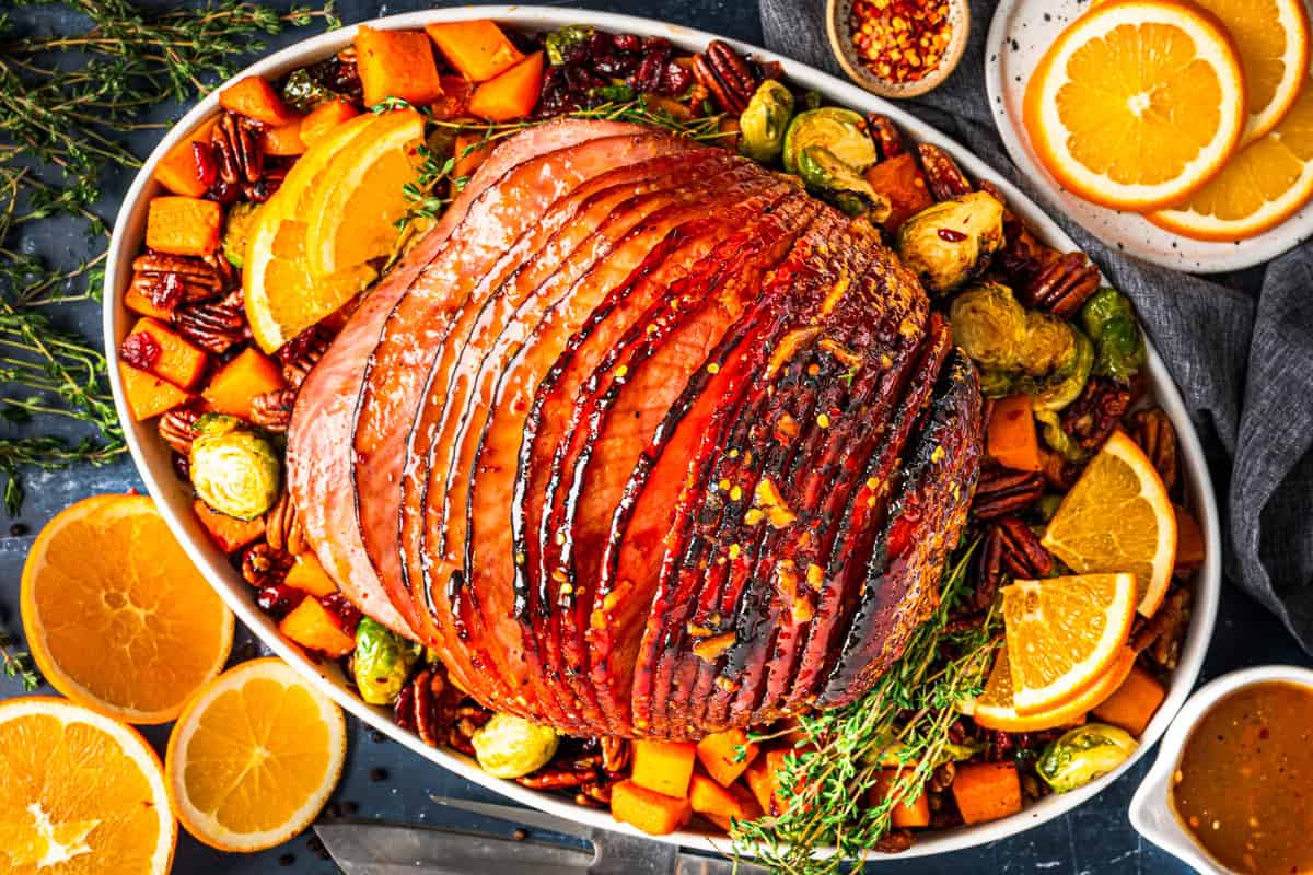 A glazed baked ham sits on a platter surrounded by roasted brussels sprouts, cubed butternut squash, pecans, and fresh herbs. Sliced oranges and a small bowl of pepper flakes are arranged nearby.