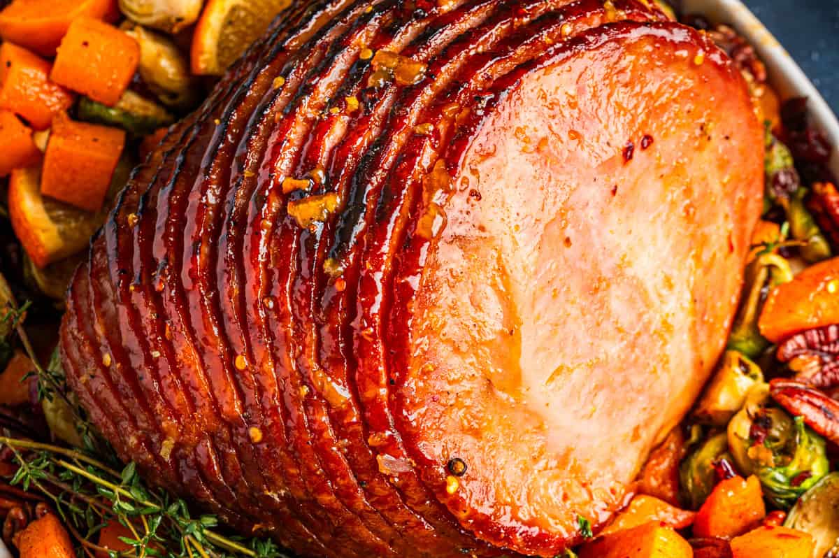 Close-up of a glazed, spiral-cut ham surrounded by roasted vegetables, including butternut squash and herbs, with a shiny, caramelized surface.