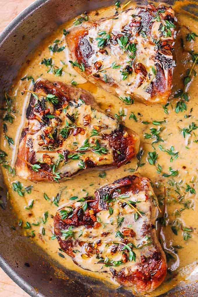 Three browned pork chops in a skillet, topped with fresh herbs and surrounded by a creamy garlic and herb sauce.