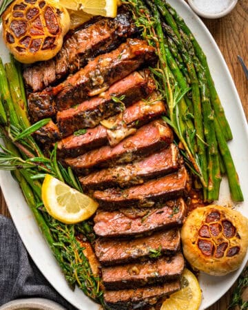 Sliced grilled steak served on a white oval plate with roasted garlic heads, fresh asparagus, sprigs of herbs, and lemon wedges, set on a wooden table with a fork and napkin nearby.
