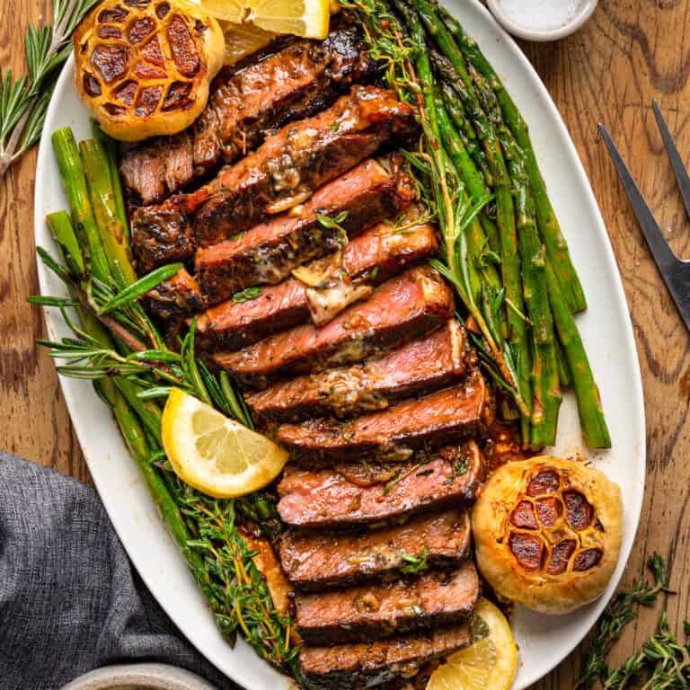 Sliced grilled steak served on a white platter with roasted garlic, asparagus, green beans, fresh herbs, and lemon wedges on a wooden table.