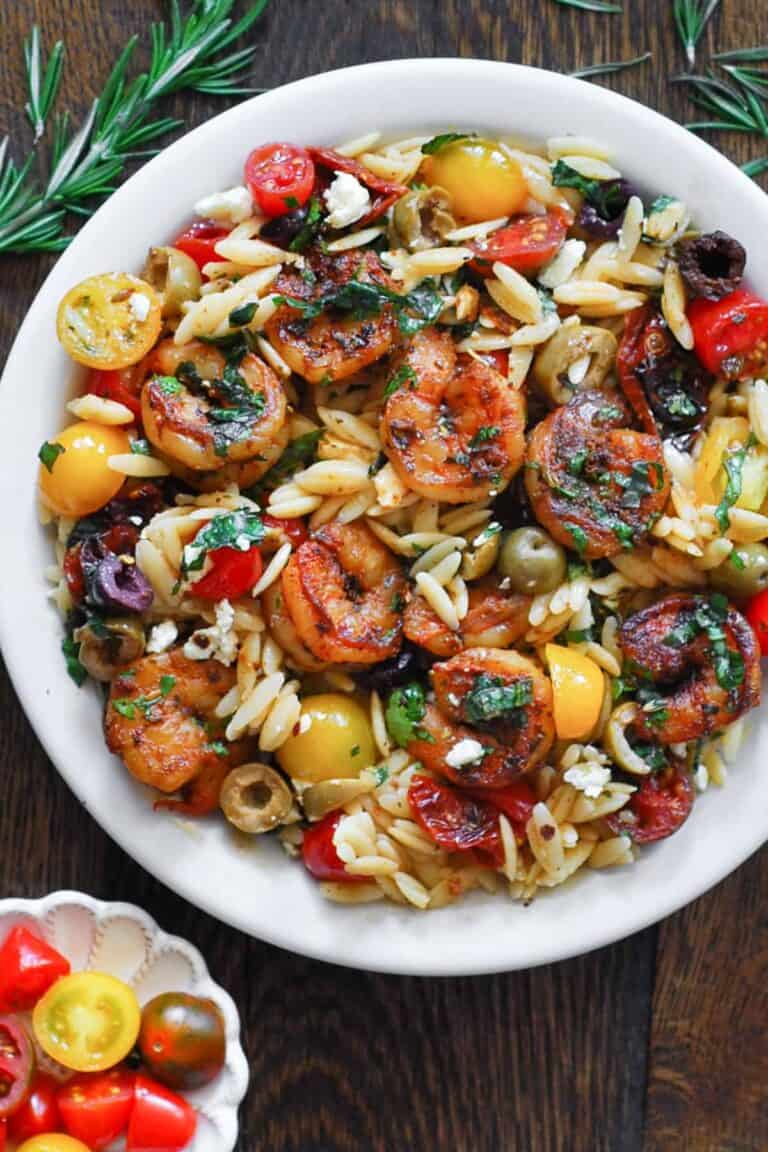 A white plate filled with orzo pasta, seared shrimp, cherry tomatoes, olives, feta cheese, and fresh herbs, served on a dark wooden table. Sprigs of rosemary and a small dish of tomatoes are nearby.