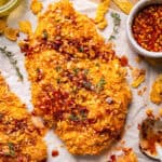 A crispy, golden breaded chicken cutlet topped with red hot honey sauce and garnished with fresh herbs, surrounded by cornflakes, a bowl of hot honey sauce, and a spoon on a parchment-lined surface.