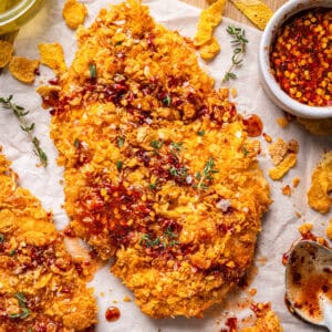 A crispy, golden breaded chicken cutlet topped with red hot honey sauce and garnished with fresh herbs, surrounded by cornflakes, a bowl of hot honey sauce, and a spoon on a parchment-lined surface.