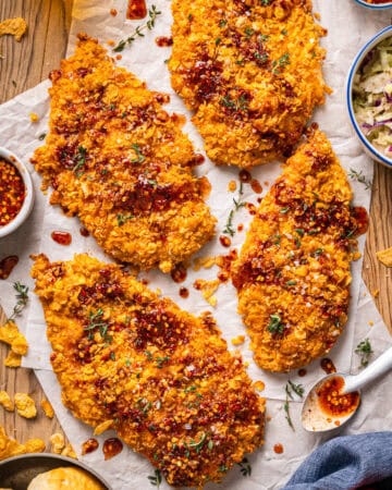 Crispy, golden baked chicken breasts drizzled with spicy hot honey sauce on parchment paper, garnished with herbs. A bowl of coleslaw and a small bowl of sauce with a spoon are beside the chicken on a wooden table.
