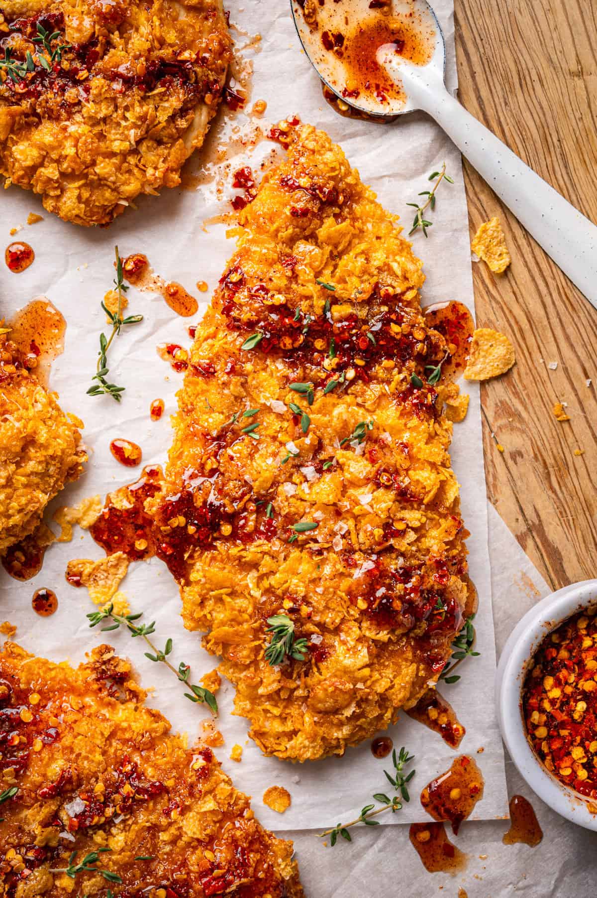 Crispy Baked Hot Honey Chicken A crispy, golden-brown breaded chicken cutlet drizzled with spicy red hot honey, garnished with fresh thyme, and surrounded by crumbs and a spoon with chili oil on parchment paper.