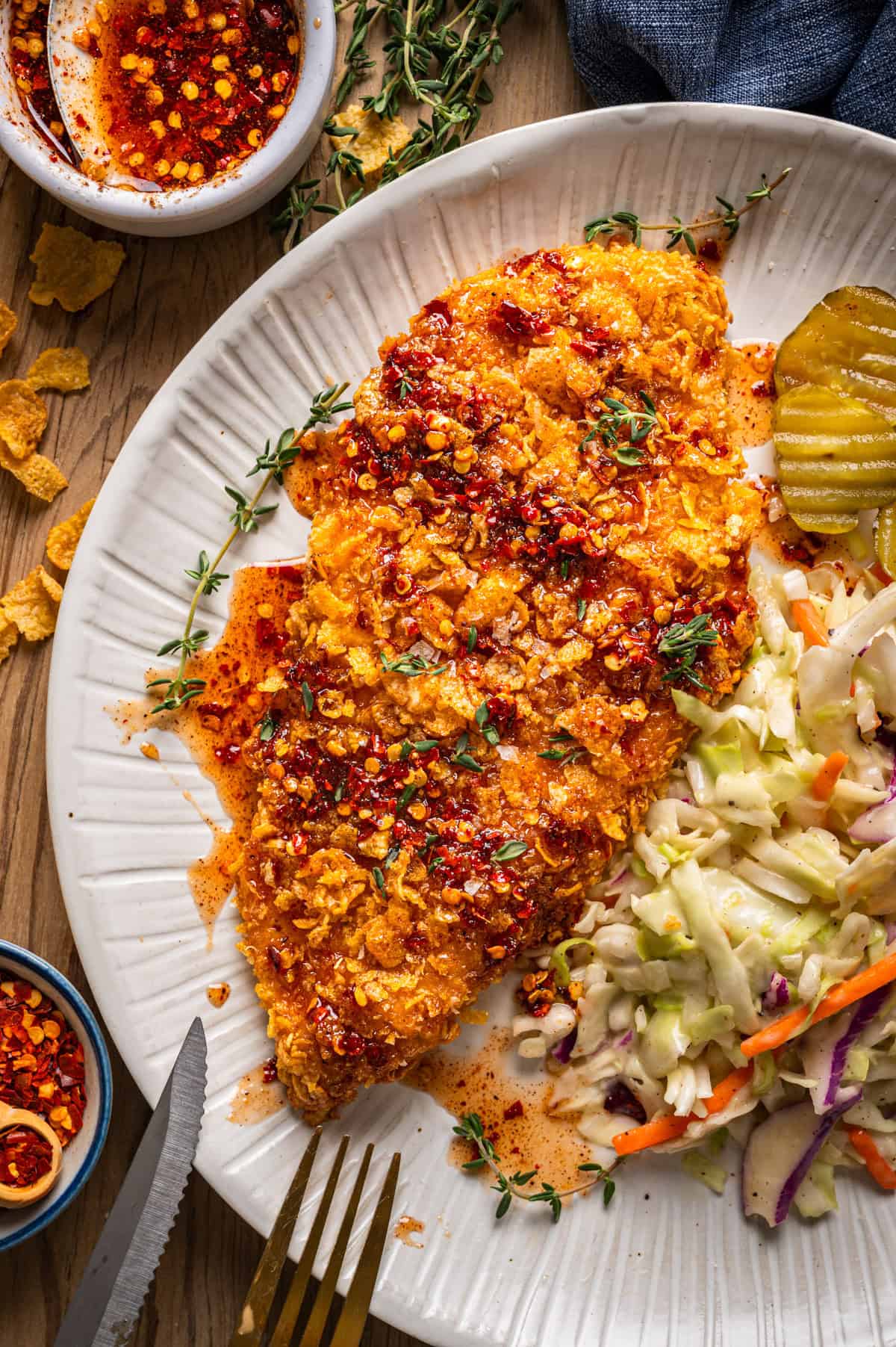 Crispy Baked Hot Honey Chicken A plate with a crispy, spicy fried chicken breast drizzled with hot honey, served with creamy coleslaw, pickle slices, and sprinkled herbs. Bowls of spicy oil and red pepper flakes are nearby.