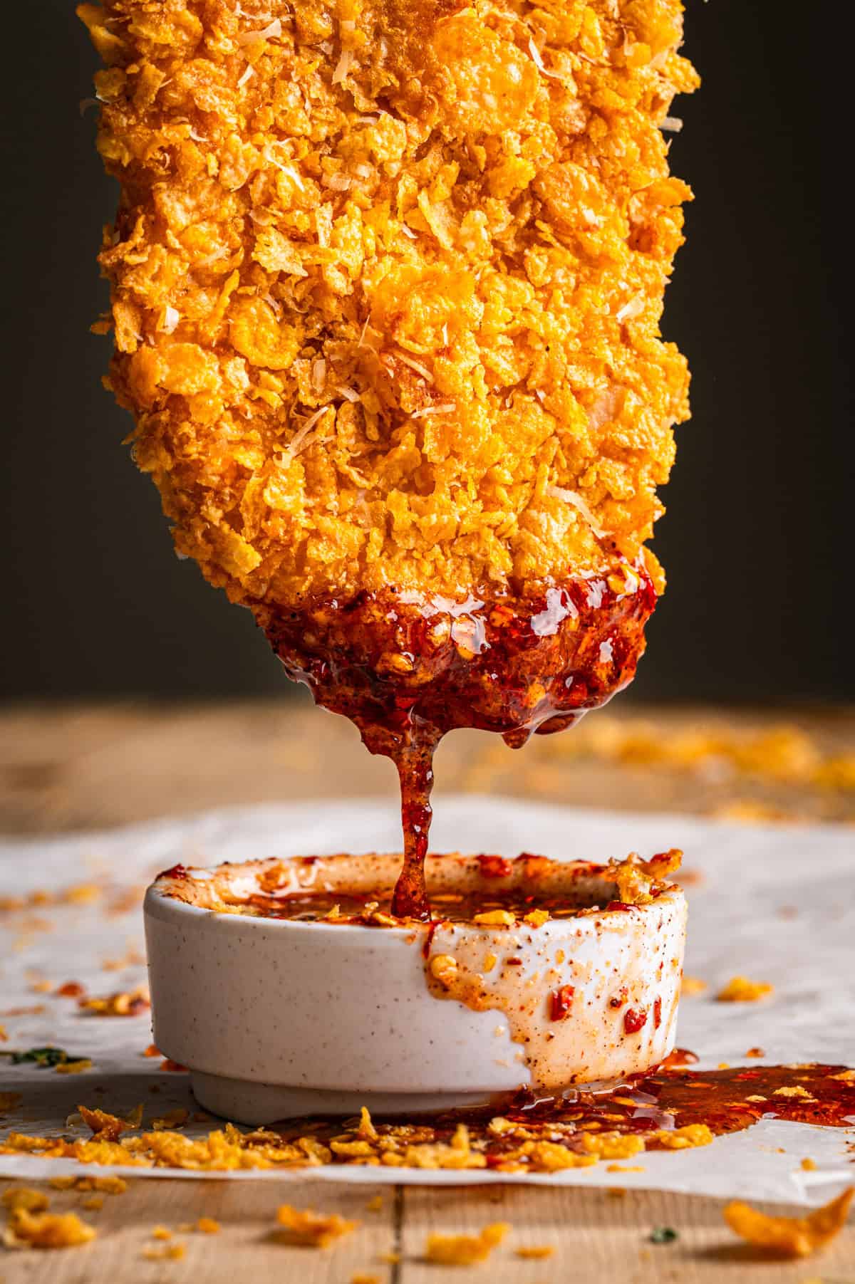 Crispy Baked Hot Honey Chicken A crispy, golden-baked chicken tender is dipped into a small bowl of spicy red hot honey sauce, with sauce dripping down and crumbs scattered around on a wooden surface.