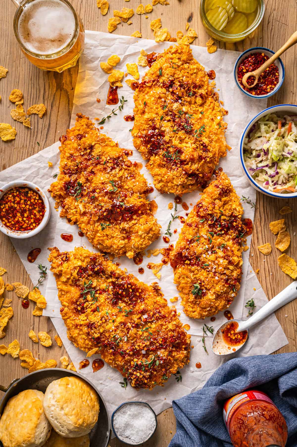 Crispy Baked Hot Honey Chicken Four crispy baked chicken breasts topped with hot honey sauce sit on parchment paper, surrounded by biscuits, coleslaw, pickles, a bowl of seasoning, and scattered cornflakes on a wooden table.