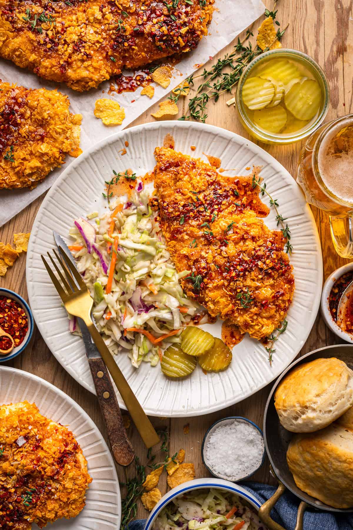 Crispy Baked Hot Honey Chicken A plate with crispy baked hot honey chicken, coleslaw, and pickle slices. Surrounding the plate are biscuits, extra fried chicken (baked), a glass of beer, pickles, and bowls of red pepper flakes, salt, and coleslaw on a wooden table.
