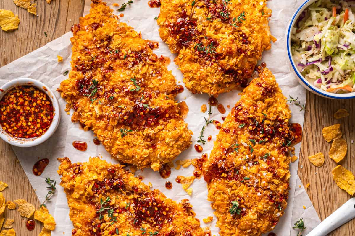 Crispy Baked Hot Honey Chicken - perfect for Game Day, Super Bowl, March Madness, or Weeknight Dinner. Four crispy golden fried chicken cutlets with a spicy red hot honey sauce drizzled on top, garnished with herbs, served on parchment paper. A bowl of coleslaw and a small bowl of chili sauce are nearby.