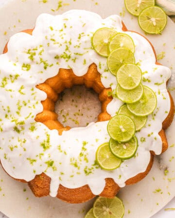 A round key lime bundt cake with white icing, topped with thin lime slices and lime zest, sits on a white surface with extra lime slices nearby.