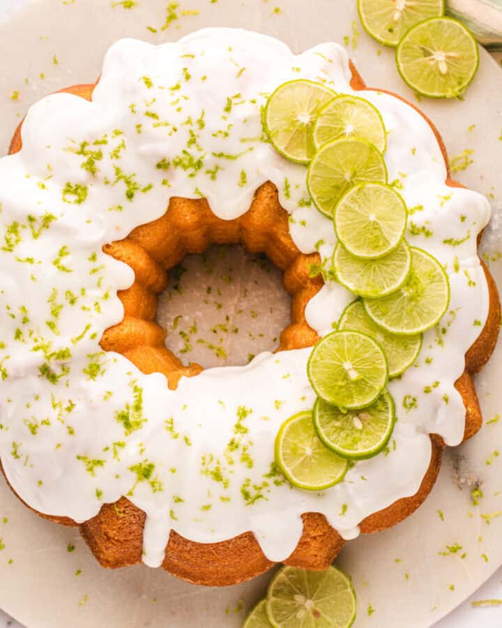A round key lime bundt cake with white icing, topped with thin lime slices and lime zest, sits on a white surface with extra lime slices nearby.