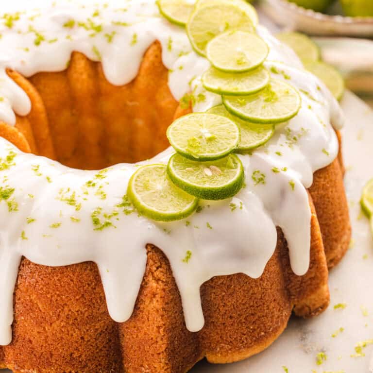 A key lime bundt cake with white icing drips, topped with slices of fresh lime and lime zest, sits on a light surface.