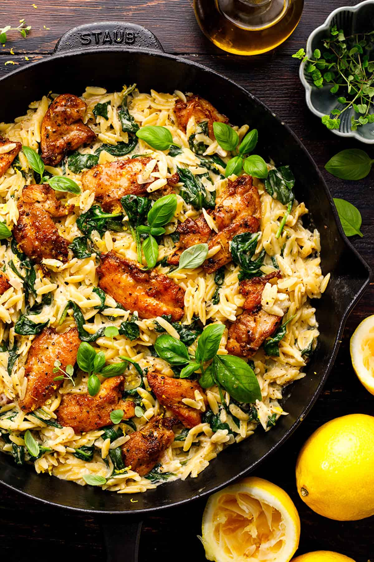 A cast iron skillet filled with creamy orzo pasta, saut&eacute;ed greens, and golden-brown chicken, garnished with fresh basil leaves. Lemons, herbs, and a bottle of oil are arranged around the skillet on a dark surface.