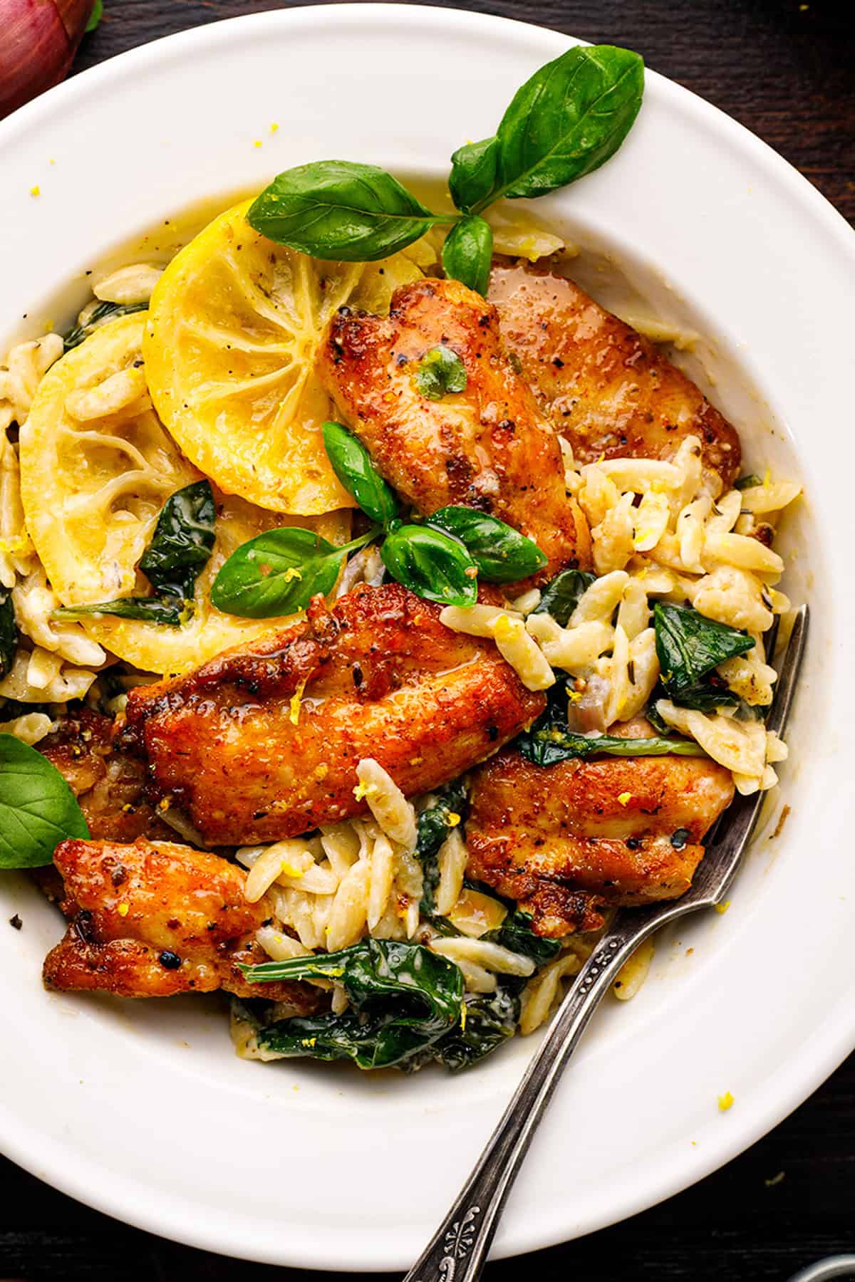 A white bowl filled with creamy chicken orzo pasta, saut&eacute;ed spinach, golden-brown chicken pieces, lemon slices, and fresh basil leaves. 