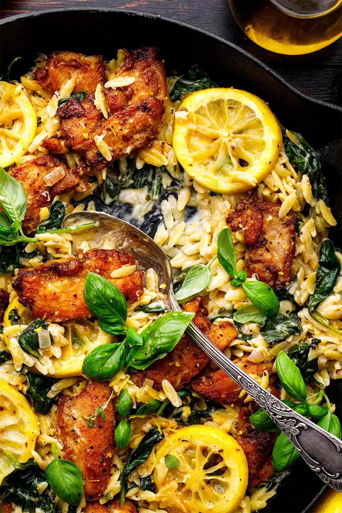 A skillet filled with creamy chicken orzo pasta, saut&eacute;ed spinach, golden-brown chicken pieces, lemon slices, and fresh basil leaves, with a spoon resting inside.