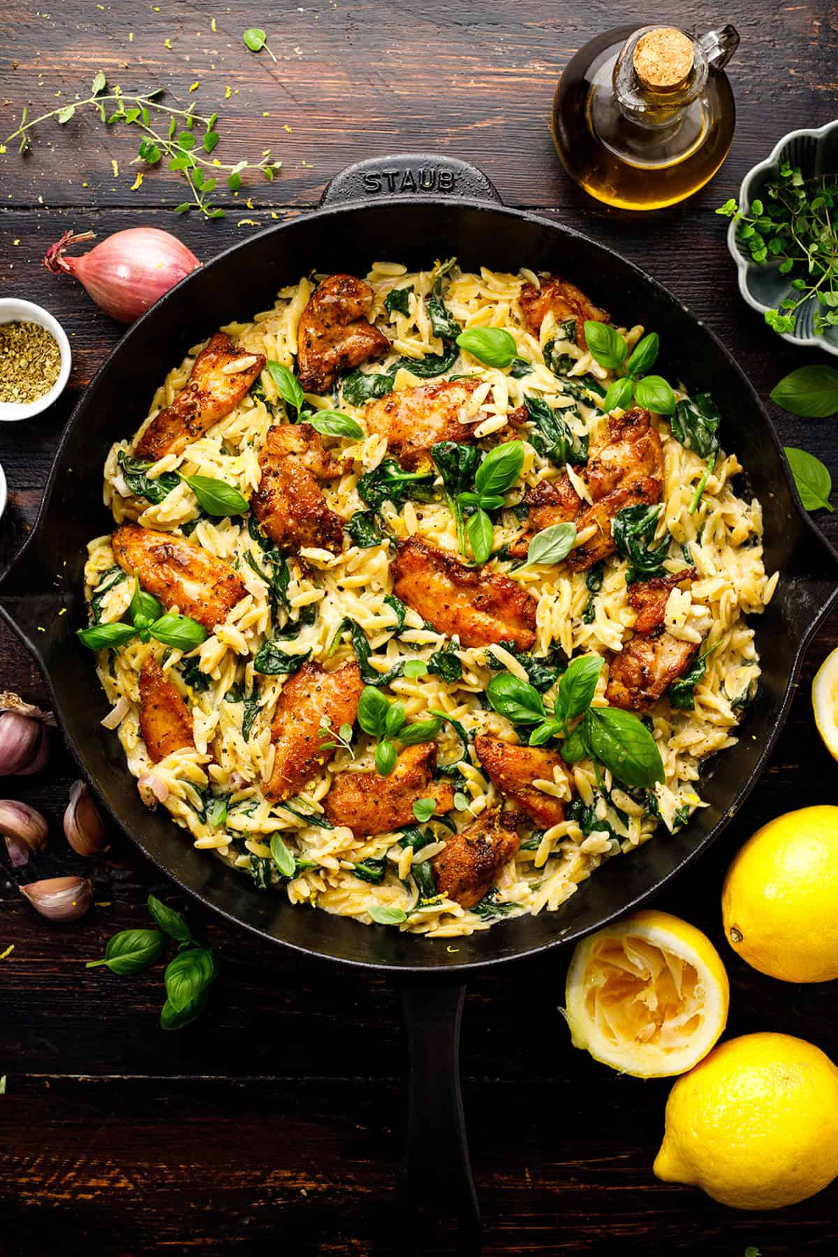 A skillet filled with creamy orzo pasta, cooked spinach, and seasoned chicken pieces, garnished with fresh basil. Surrounding the skillet are lemons, herbs, garlic, a shallot, a small bowl of spices, and a bottle of olive oil.