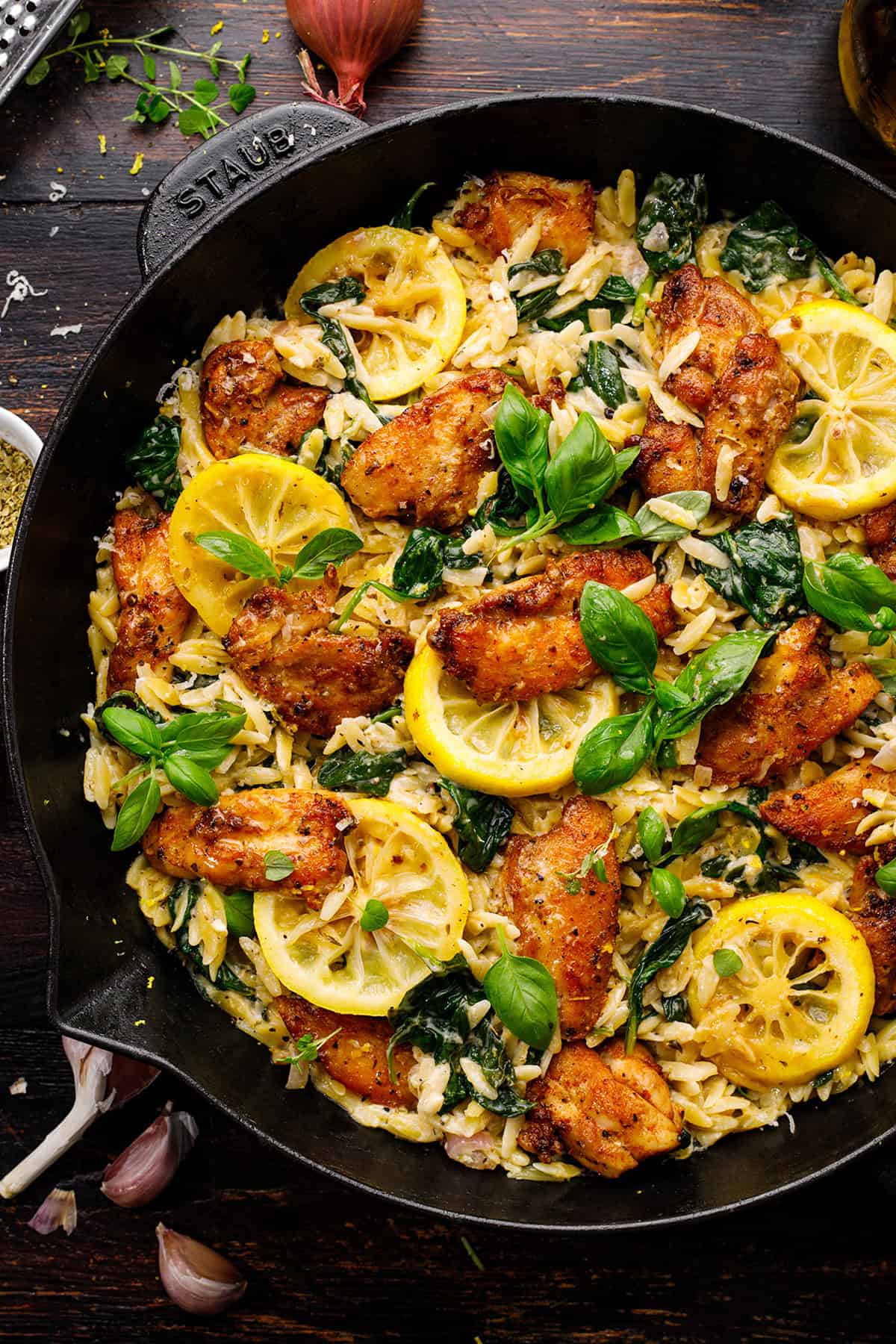 A skillet filled with creamy chicken orzo pasta, saut&eacute;ed spinach, seared chicken pieces, lemon slices, and fresh basil leaves on top, arranged on a dark wooden surface.