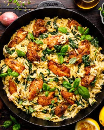 A skillet filled with creamy lemon chicken orzo pasta, saut&eacute;ed spinach, and seasoned chicken pieces, garnished with fresh basil leaves. Surrounding the skillet are herbs, lemon halves, garlic, shallots, and a small bowl of spices.