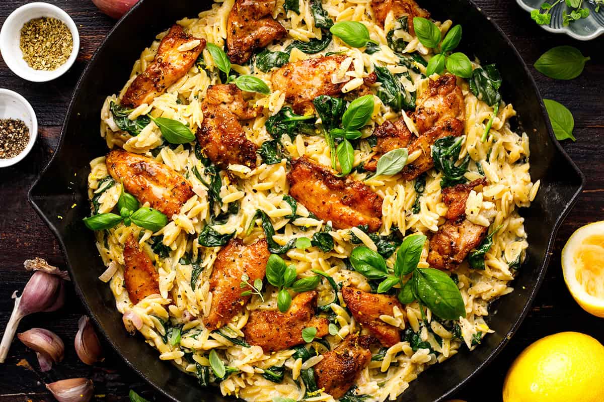 A skillet filled with creamy chicken orzo pasta, saut&eacute;ed spinach, and seasoned chicken pieces, garnished with fresh basil leaves. Garlic cloves, lemon halves, and small bowls of spices are visible nearby.
