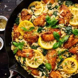 A skillet filled with lemon chicken orzo pasta, seasoned chicken pieces, spinach, lemon slices, and fresh basil leaves, garnished with herbs.