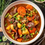 A bowl of Mulligan beef stew, filled with chunks of beef, carrots, potatoes, green beans, peas, corn, and mushrooms in a rich broth, garnished with fresh herbs.