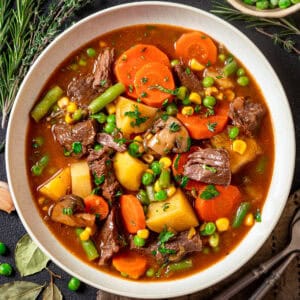A bowl of Mulligan beef stew, filled with chunks of beef, carrots, potatoes, green beans, peas, corn, and mushrooms in a rich broth, garnished with fresh herbs.