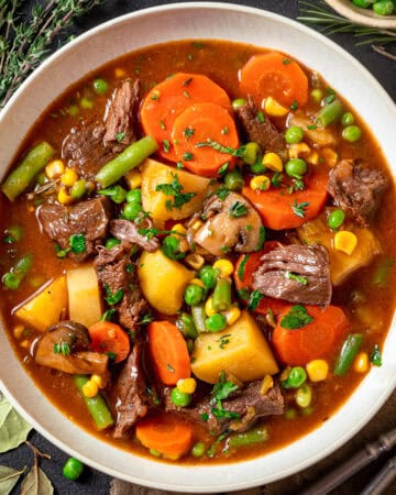A bowl of Mulligan beef stew, filled with chunks of beef, carrots, potatoes, green beans, peas, corn, and mushrooms in a rich broth, garnished with fresh herbs.