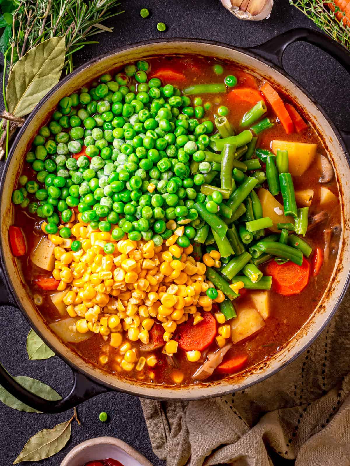 Mulligan Stew (Hobo Stew) A pot of hearty mulligan stew with visible peas, green beans, corn, carrots, and potatoes in a rich broth, surrounded by fresh herbs and a green kitchen towel.
