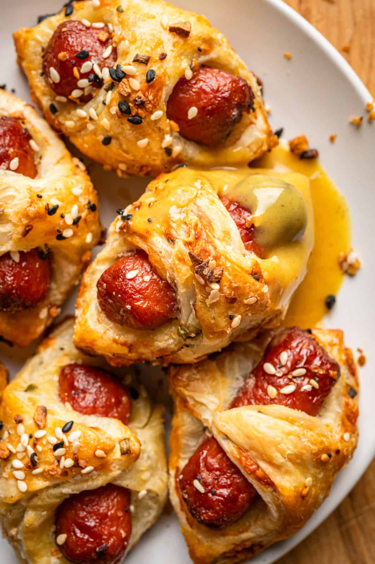 Pigs in a Blanket - Golden, flaky pastry wraps mini sausages, sprinkled with mixed seeds, on a white plate. One pastry oozes melted cheese, and crumbs are scattered around, showing a fresh, savory snack.