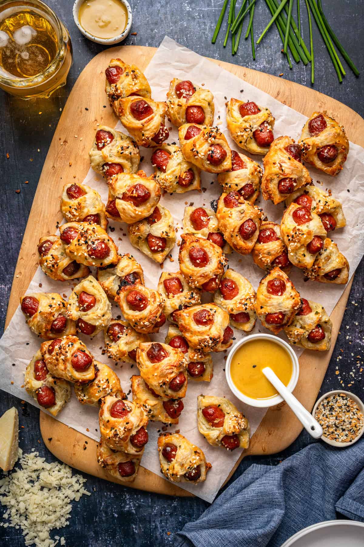 A wooden board holds Pigs in a Blanket - mini puff pastry snacks filled with sausage and topped with sesame seeds, arranged on parchment paper. Nearby are dipping sauces, shredded cheese, chives, and a glass of beer.
