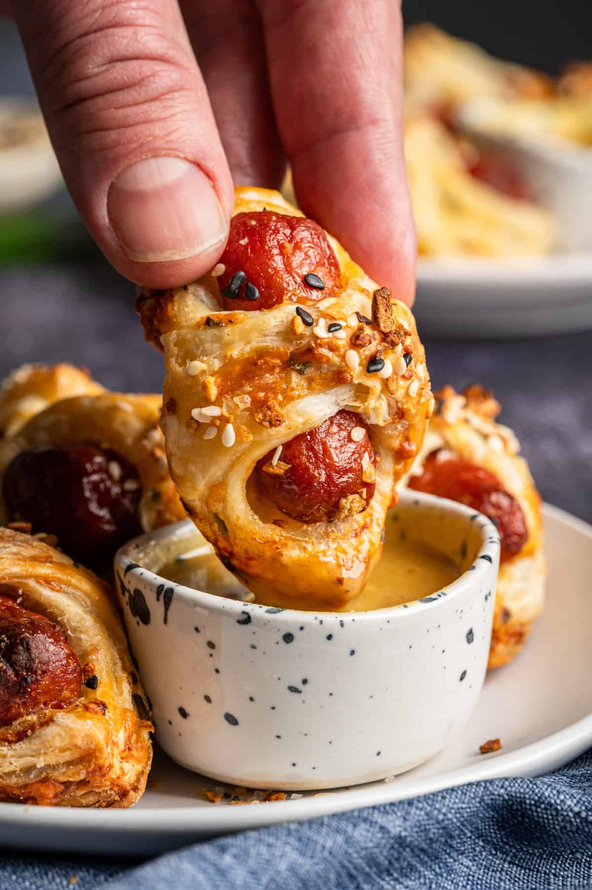 A hand dips a puff pastry-wrapped sausage (Pigs in a Blanket) topped with seeds into a small speckled bowl of mustard sauce, with more pastry-wrapped sausages on a plate in the background.