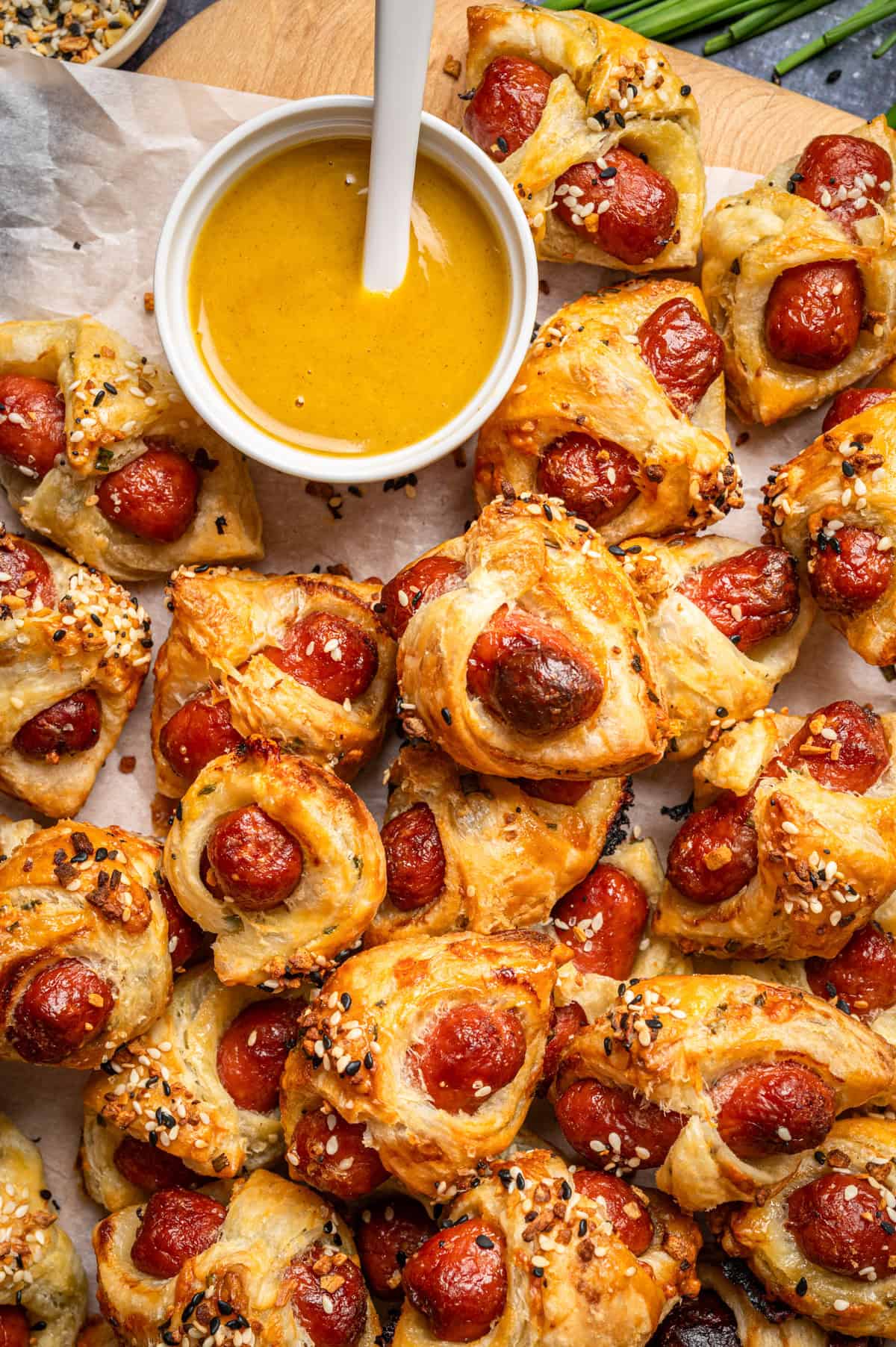 A pile of puff pastry-wrapped mini sausages (Pigs in a Blanket) sprinkled with Everything Bagel seasoning, arranged on parchment paper next to a bowl of yellow dipping sauce with a white spoon.