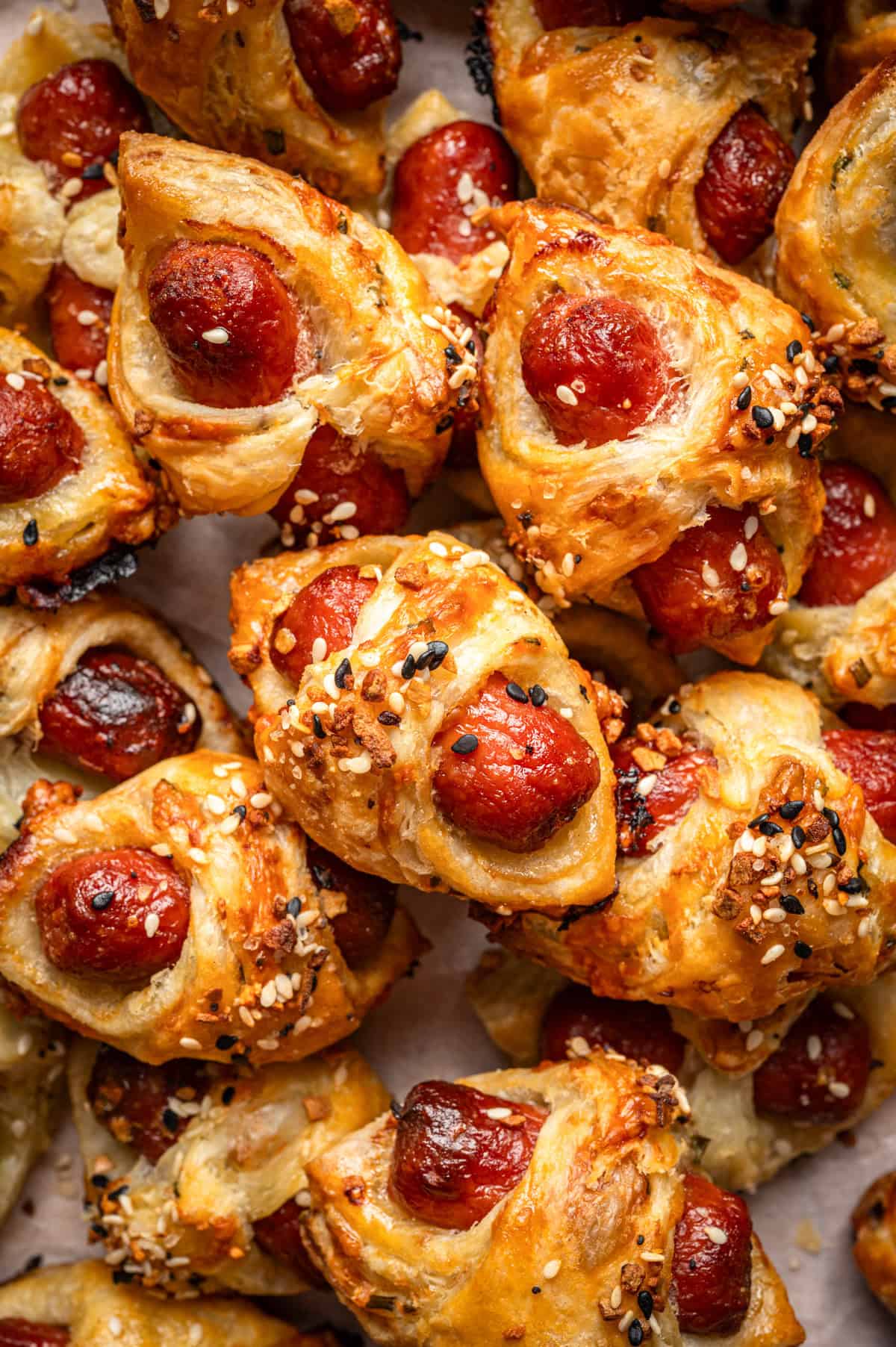 Close-up of golden-brown pigs in a blanket: mini sausages wrapped in flaky puff pastry, topped with sesame and poppy seeds, baked and piled together on a tray.