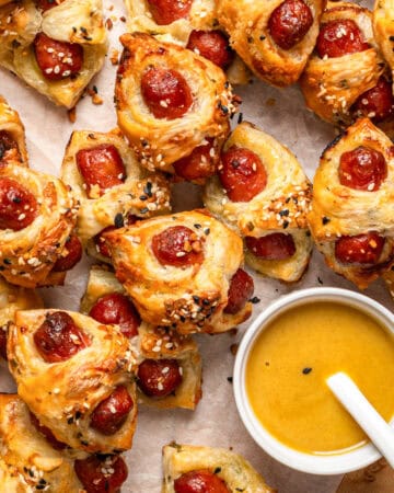 Golden-brown puff pastry bites (Pigs in a Blanket) filled with mini sausages, sprinkled with sesame and poppy seeds (Everything Bagel Seasoning), arranged on parchment paper next to a bowl of yellow dipping sauce with a white spoon.