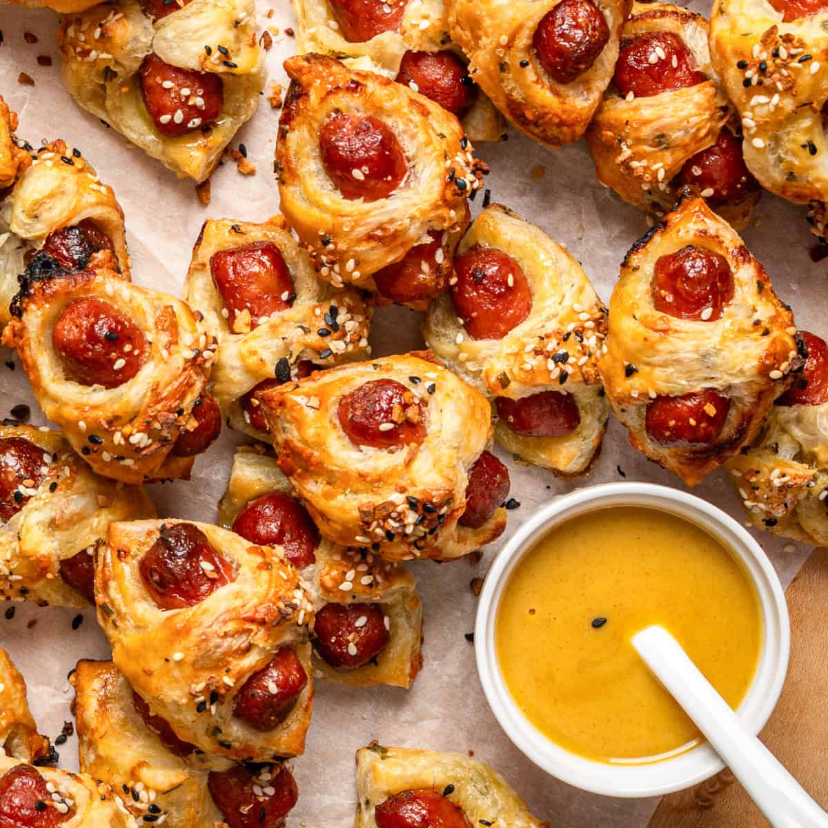 Golden-brown puff pastry bites (Pigs in a Blanket) filled with mini sausages, sprinkled with sesame and poppy seeds (Everything Bagel Seasoning), arranged on parchment paper next to a bowl of yellow dipping sauce with a white spoon.