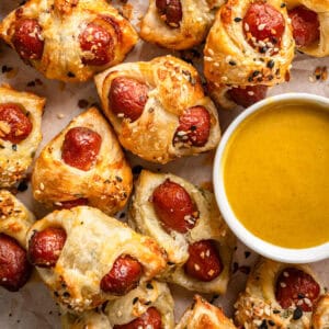 A close-up of pigs in a blanket with golden, flaky pastry, sprinkled with Everything Bagel seasoning (sesame seeds), arranged around a bowl of yellow dipping sauce on parchment paper.