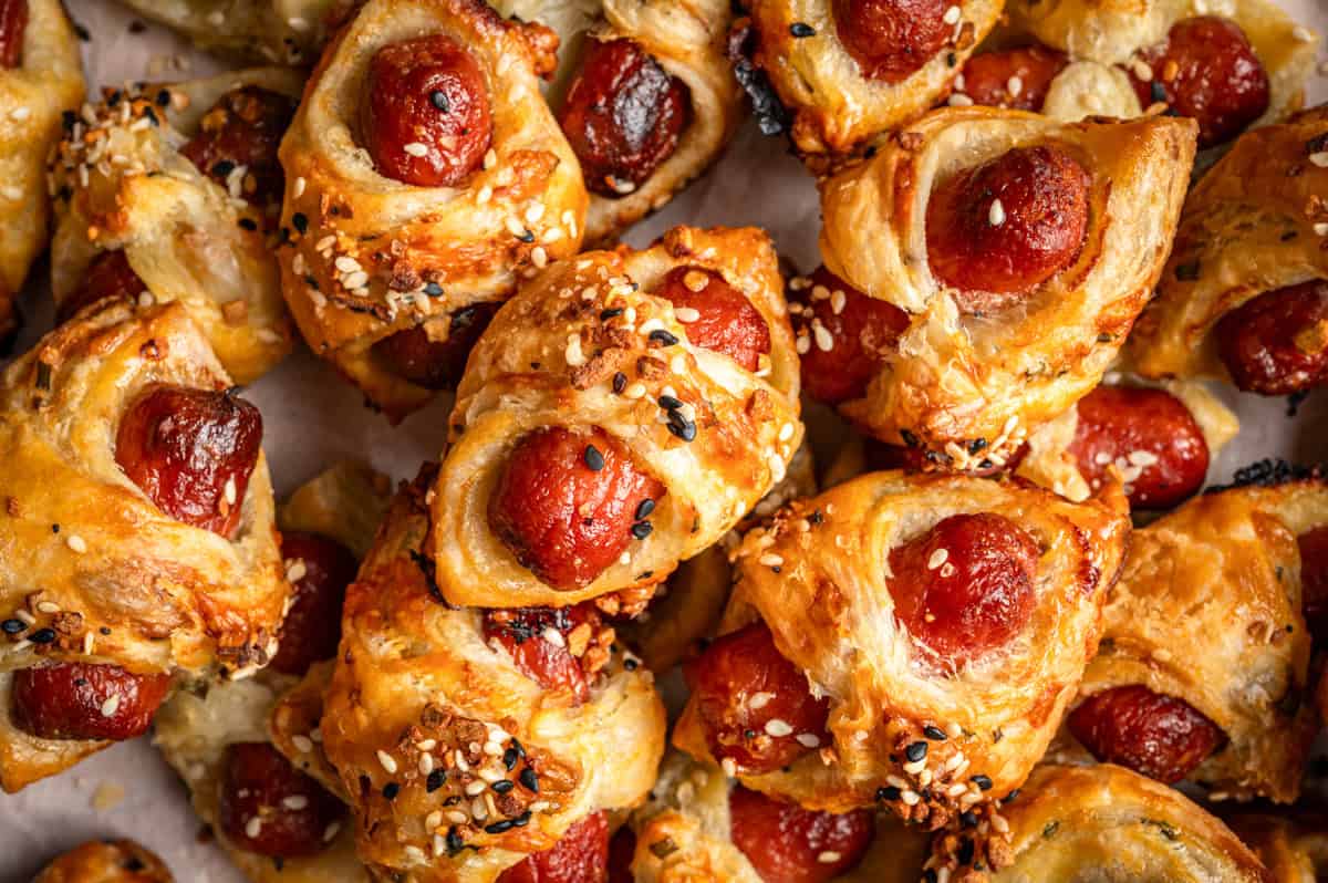 A close-up of golden-brown puff pastry pigs in a blanket wrapped around mini sausages, topped with a sprinkle of sesame and poppy seeds. 