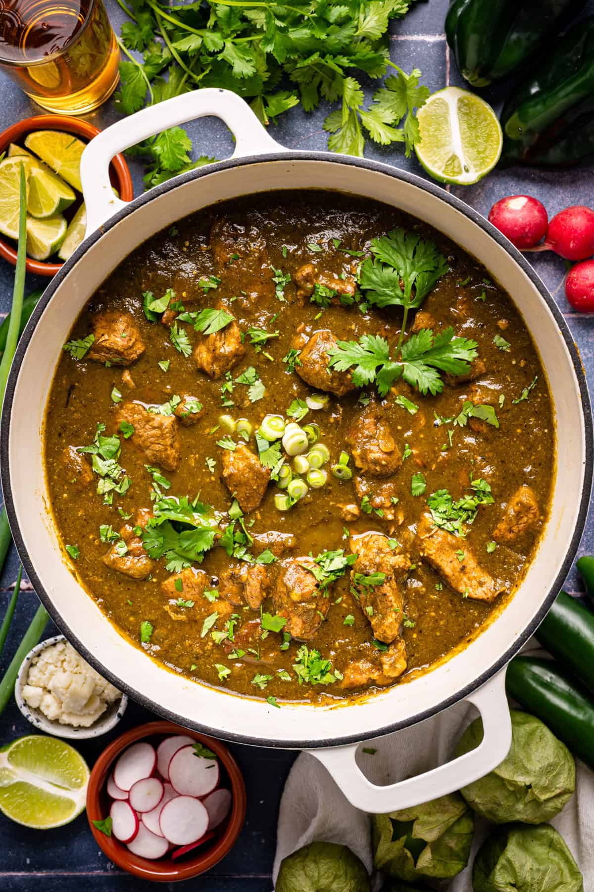 A pot of green chile pork stew garnished with cilantro and sliced green onions, surrounded by fresh limes, radishes, tomatillos, cilantro, jalape&ntilde;os, and small bowls of toppings on a dark surface.