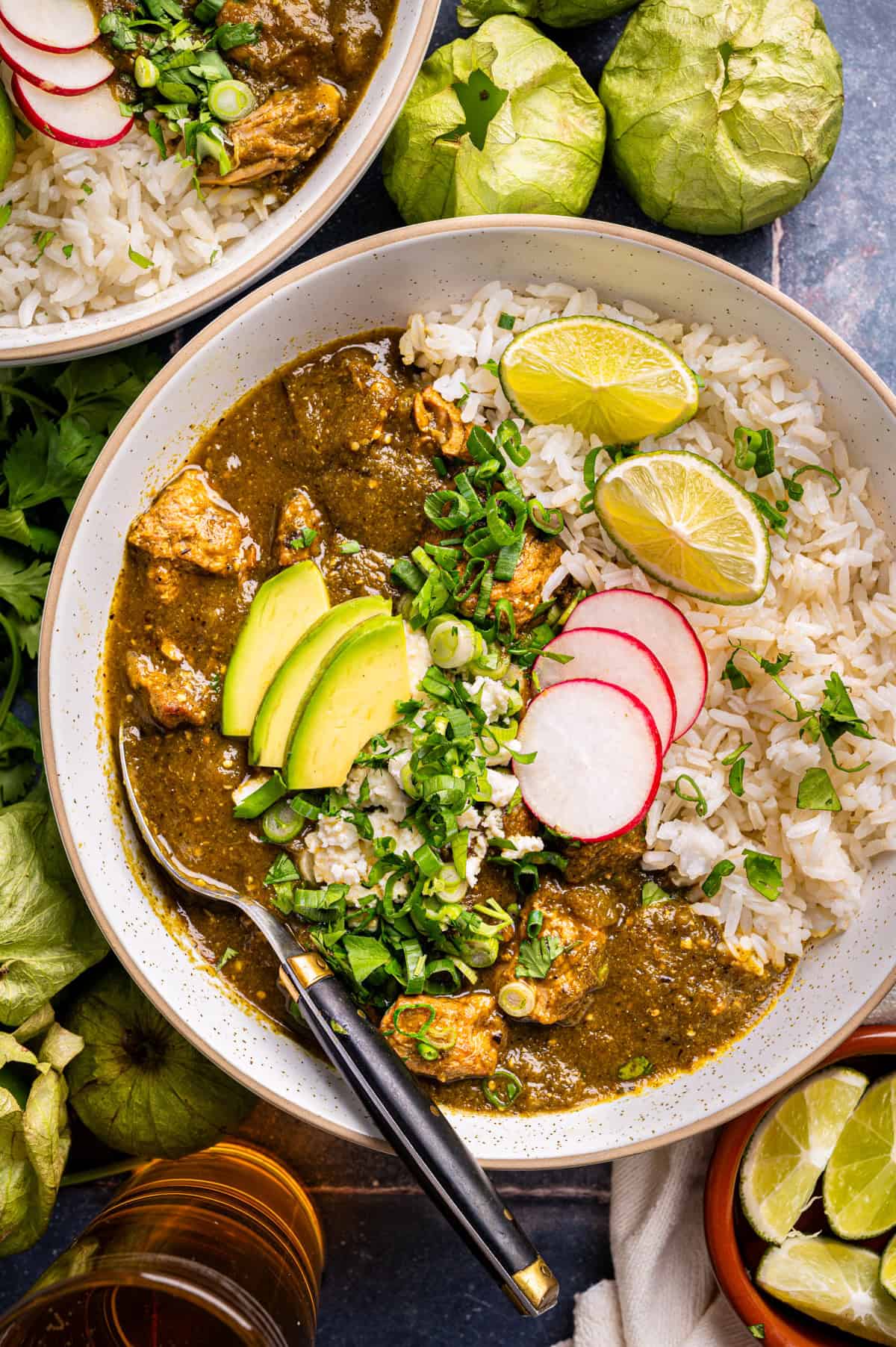 A bowl of green chili stew with chunks of pork, served with white rice, sliced avocado, radishes, green onions, cilantro, and lime wedges. Tomatillos, fresh herbs, and another bowl of stew are nearby.