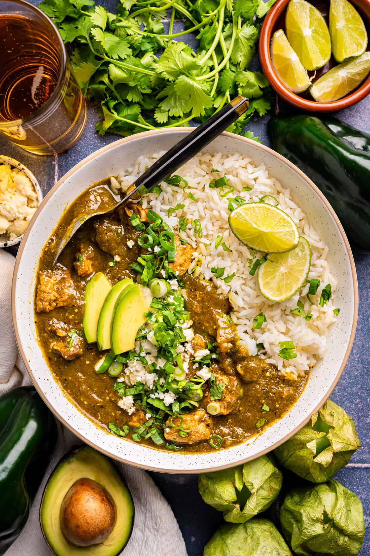 A bowl of white rice and pork chile verde (green chile stew) garnished with lime wedges, avocado slices, chopped green onions, and cilantro. Fresh tomatillos, peppers, avocado, lime wedges, and herbs are arranged around the bowl.