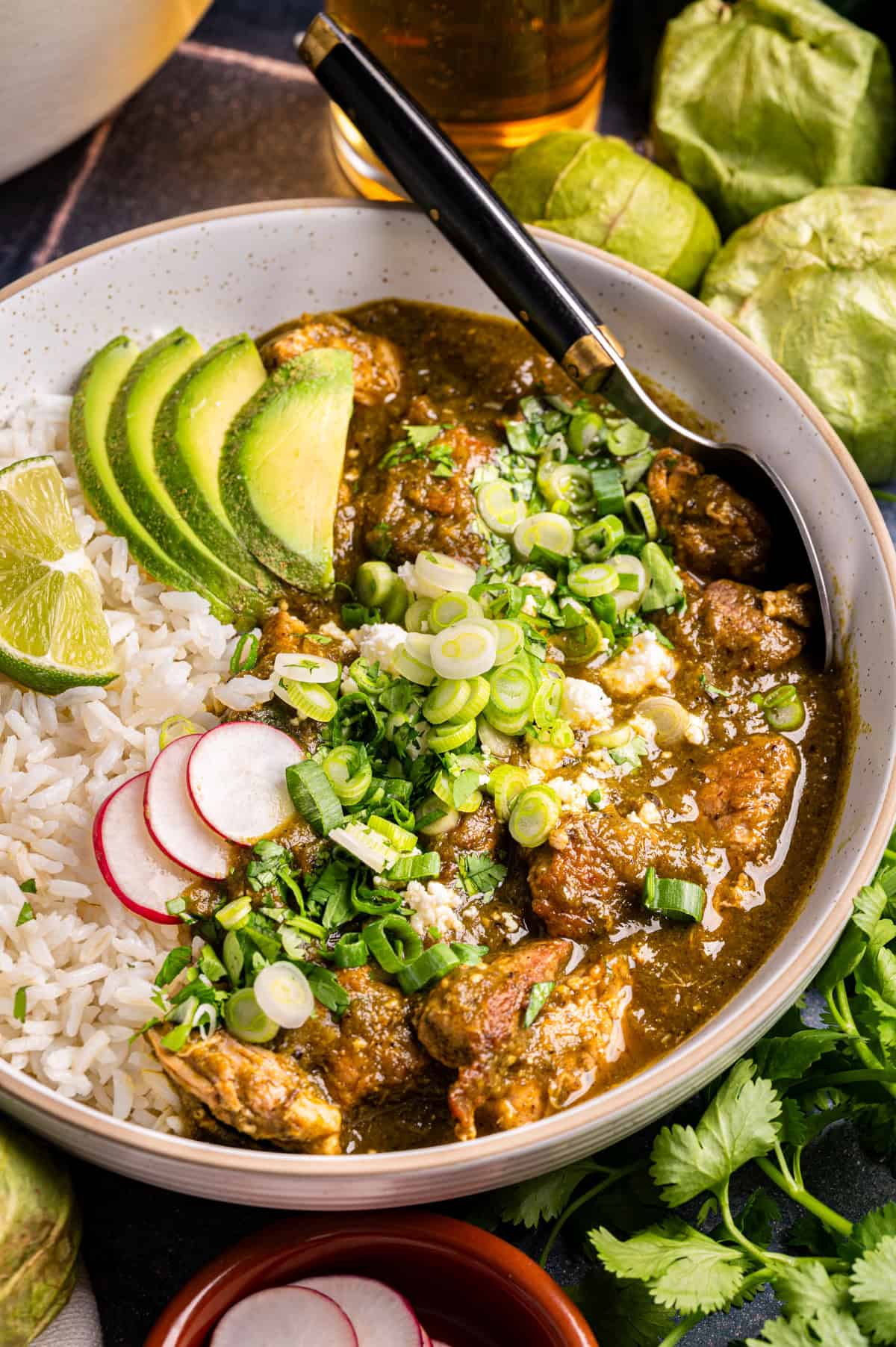 A bowl of Mexican green chile pork stew with white rice, garnished with sliced avocado, radishes, lime wedges, green onions, cilantro, and cheese; a spoon rests in the bowl. Tomatillos and herbs are nearby.