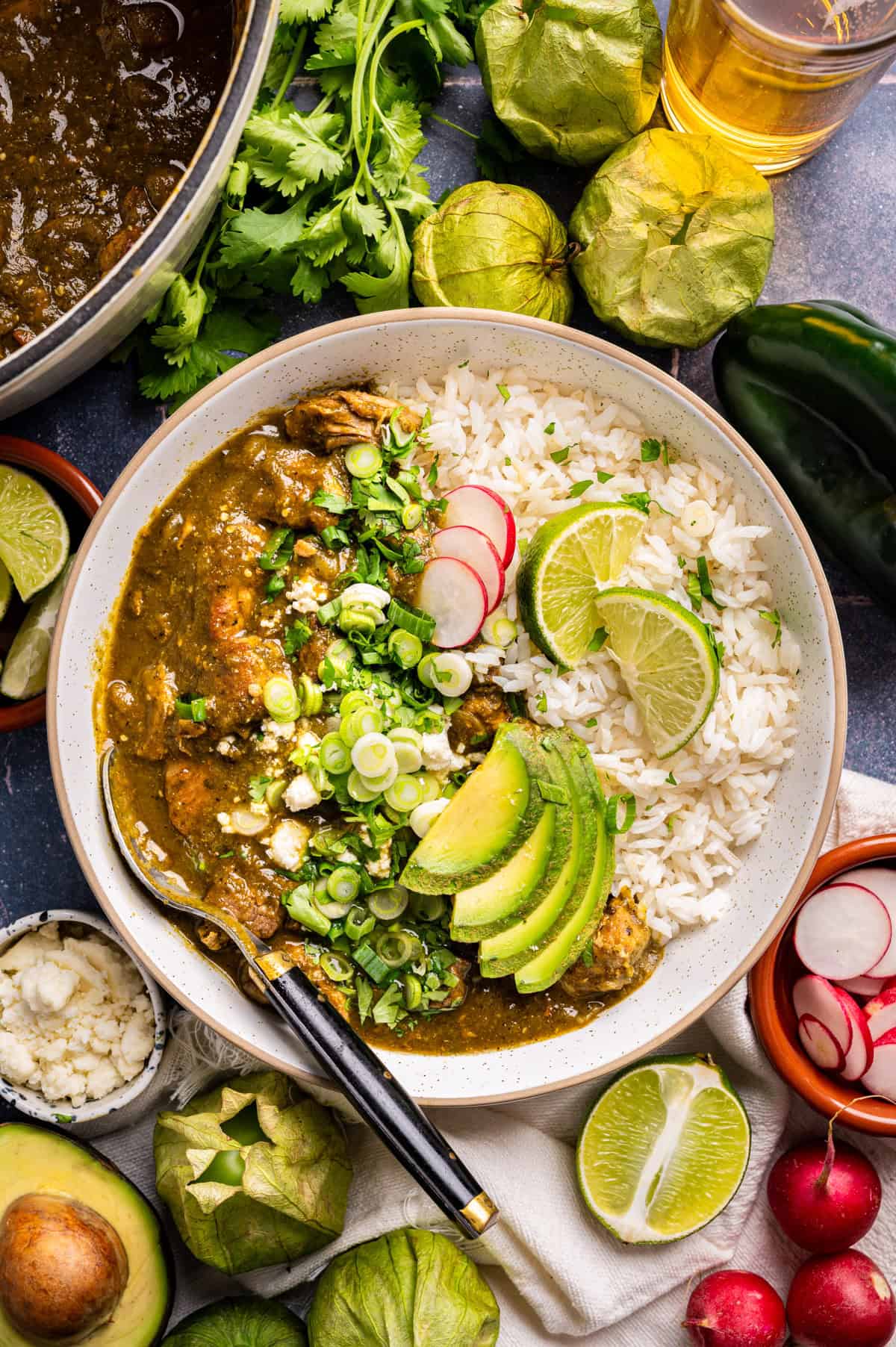 A bowl of white rice and green chili stew (pork chile verde) garnished with sliced avocado, radishes, lime wedges, green onions, and cilantro, surrounded by fresh ingredients like tomatillos, limes, avocado, radishes, and cilantro.